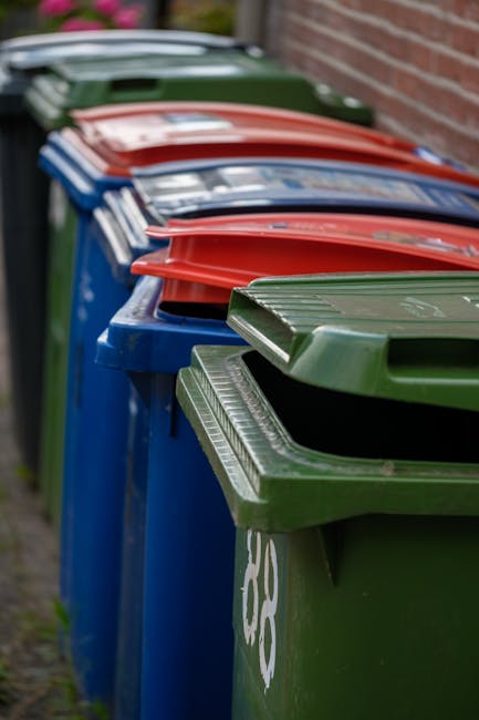 A large collection of mixed waste, consisting of various cardboard boxes, plastic bags, and paper products, is piled haphazardly on a paved area near a curb in front of a commercial building. The wast