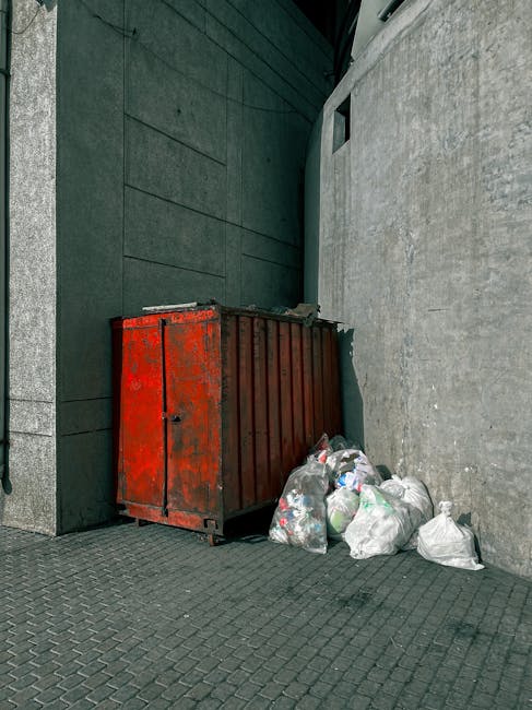 An aerial view of a large outdoor area filled with a significant amount of mixed waste and rubbish, including black and colored plastic bags, cardboard boxes, broken furniture, and discarded miscellan