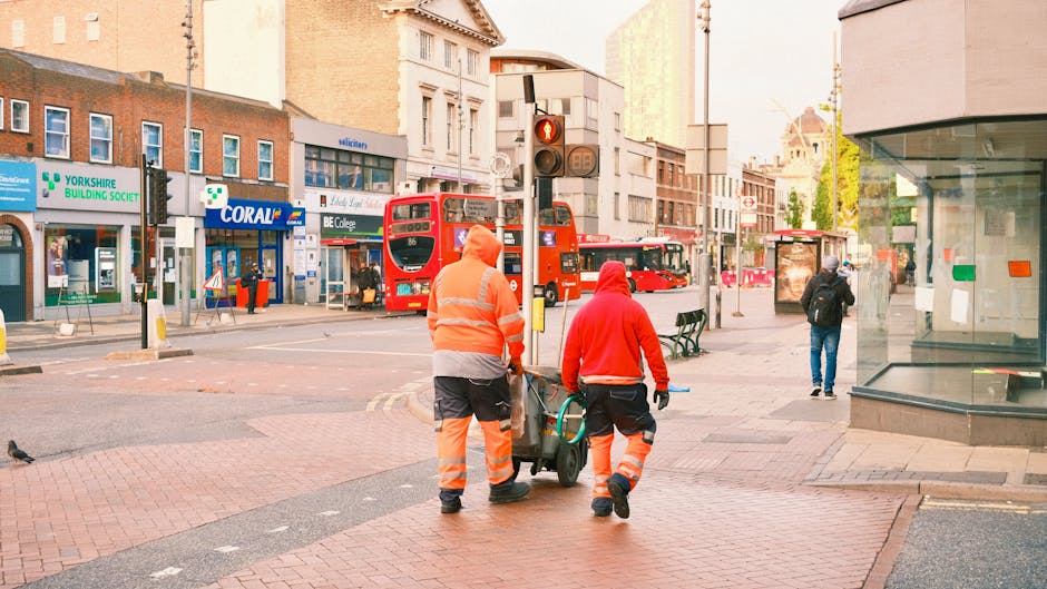 Two waste collection workers dressed in orange high-visibility safety clothing are walking across a busy urban street while pushing a wheeled bin filled with collected rubbish. The worker on the left
