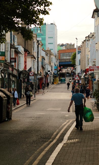 Two waste collection workers dressed in orange high-visibility safety clothing are walking across a busy urban street while pushing a wheeled bin filled with collected rubbish. The worker on the left
